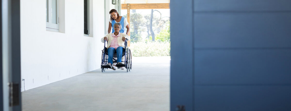 A smiling woman pushing a person in a wheelchair along a walkway beside a house
