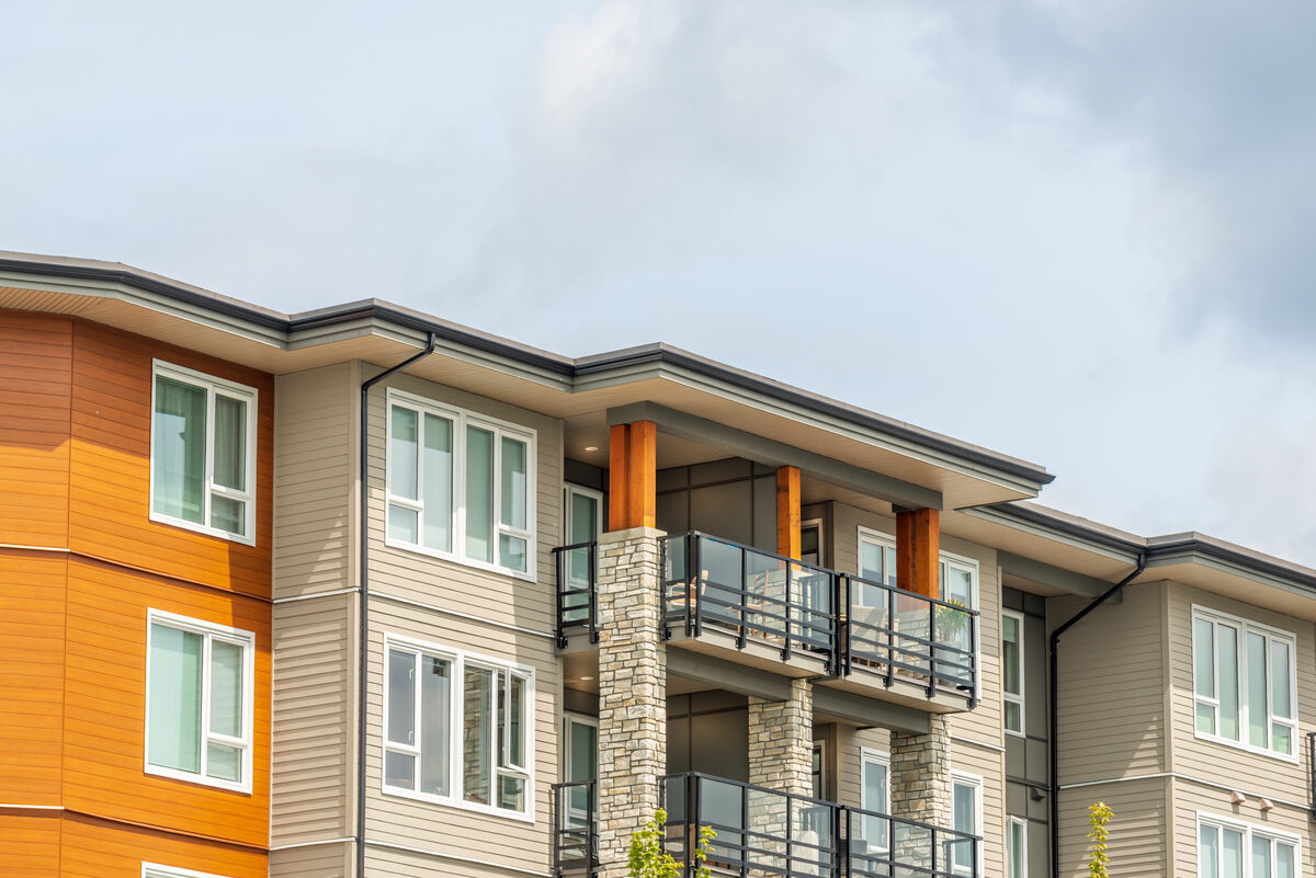 A three-story residential building with glass-railed balconies, tan and orange siding and stone-clad columns, under a pale overcast sky