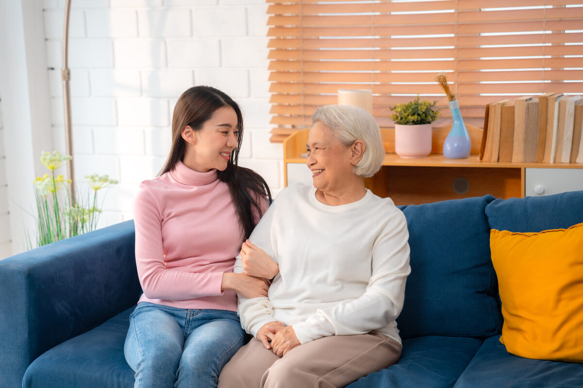 A younger woman and an older woman sitting together on a blue sofa in a bright living room, turned towards each other and smiling