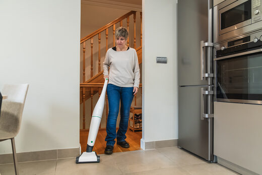 A woman using an upright vacuum in front of a wooden staircase, with a kitchen oven to one side