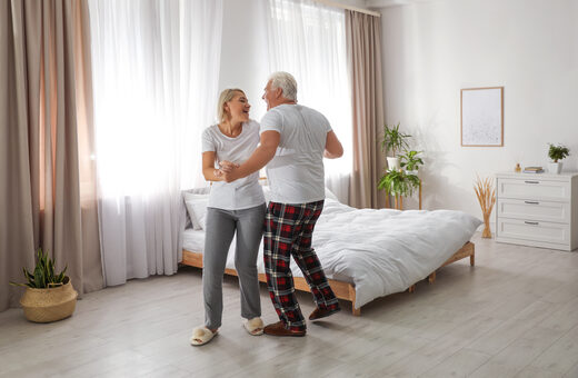 An older couple dancing together in a bright bedroom beside a low bed, with sheer curtains and a clear wood floor