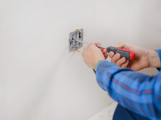 An electrician in a blue plaid shirt working on an open wall outlet with a red-handled screwdriver