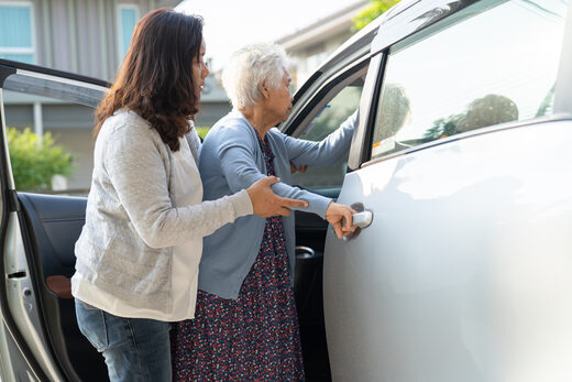 A woman holding an older woman by the arm as she opens the door of a silver car parked outside a home