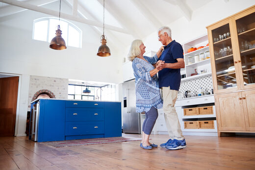 An older couple dancing in a spacious kitchen with a blue island, glass-front cabinets and a wide wood floor