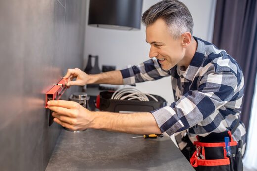 A man in a plaid shirt and tool belt holding a red spirit level against a gray kitchen wall